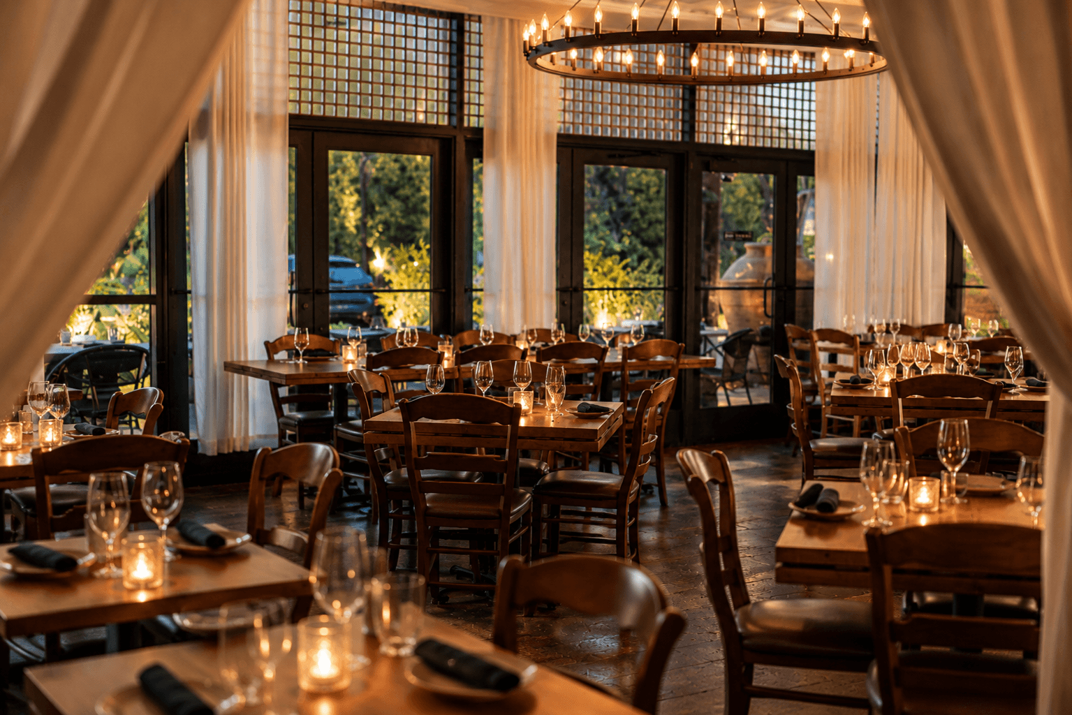 Cozy restaurant interior with wooden tables set for dining, dim lighting, and large windows revealing an outdoor view at dusk.