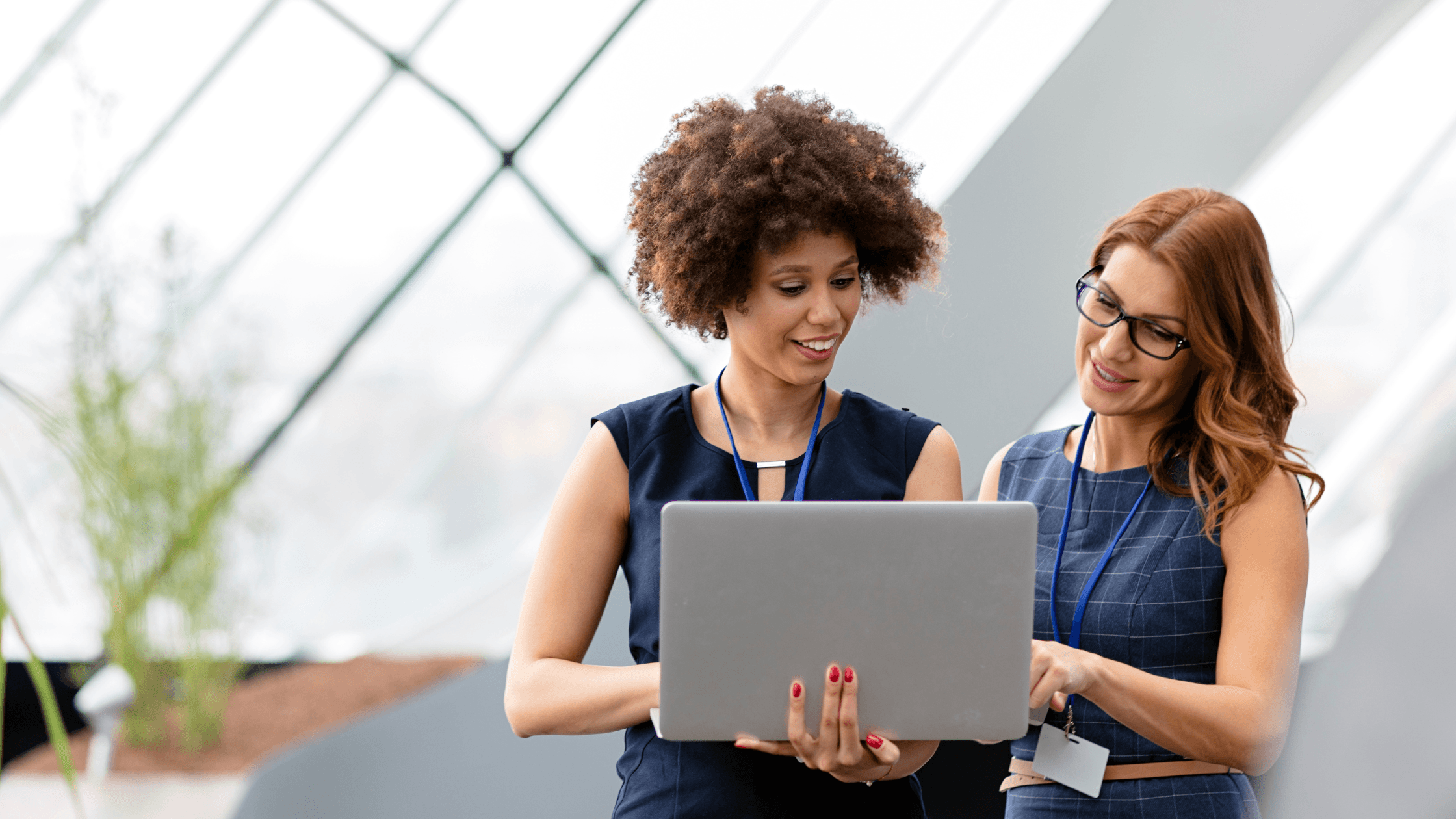 Two women smiling and discussing something on a laptop, standing in a bright, modern office with large windows.