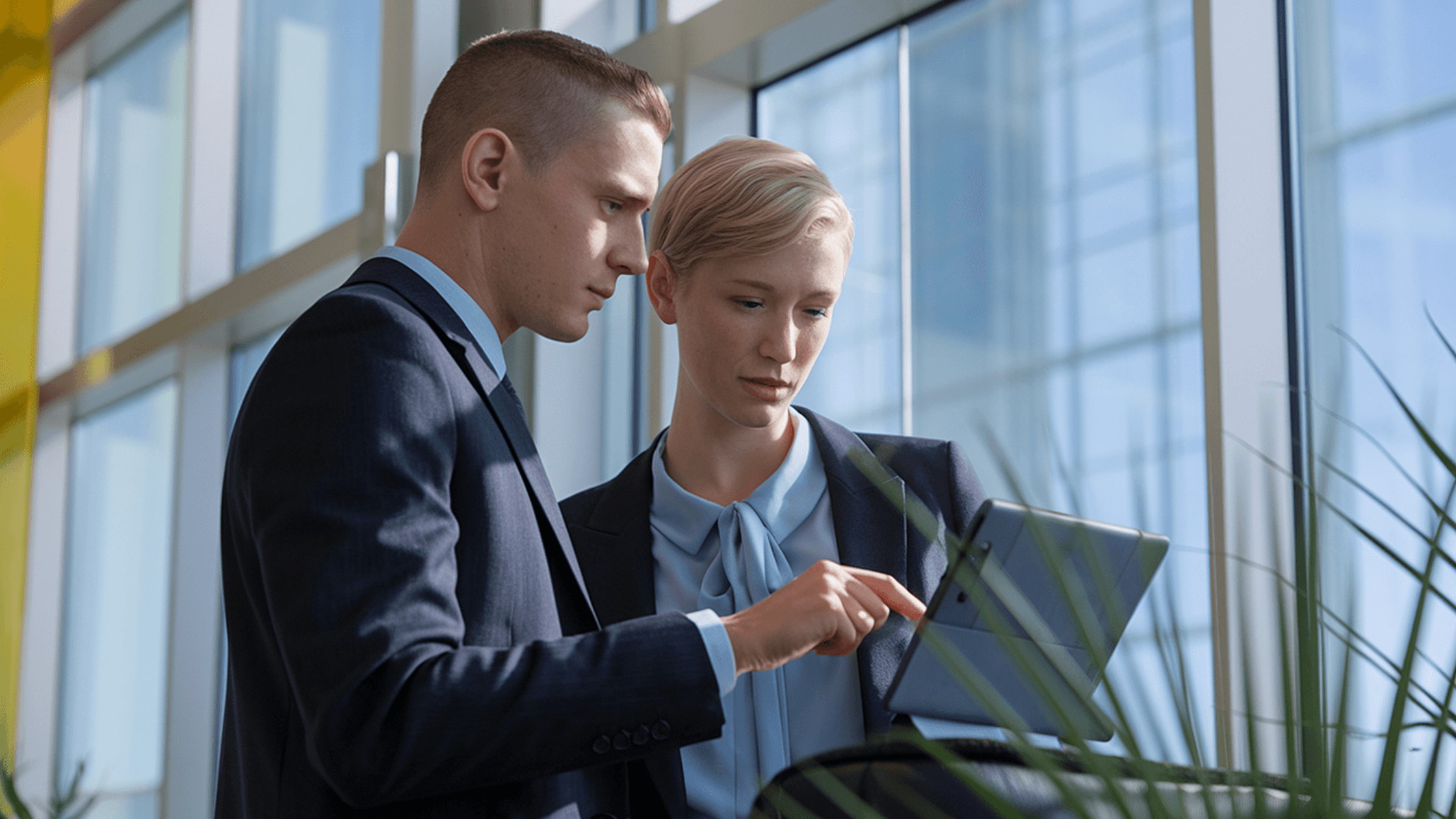 Two professionals in suits focus on a tablet screen, discussing content in a bright, modern office setting.