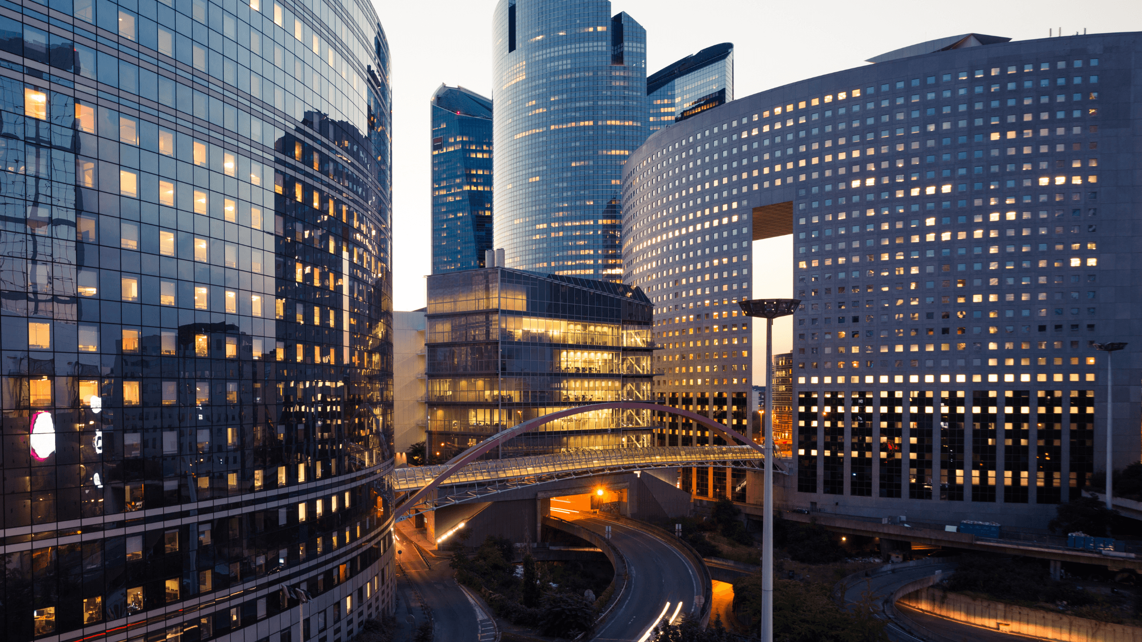 Modern cityscape at dusk with illuminated skyscrapers, a curved pedestrian bridge, and winding roads in an urban setting.