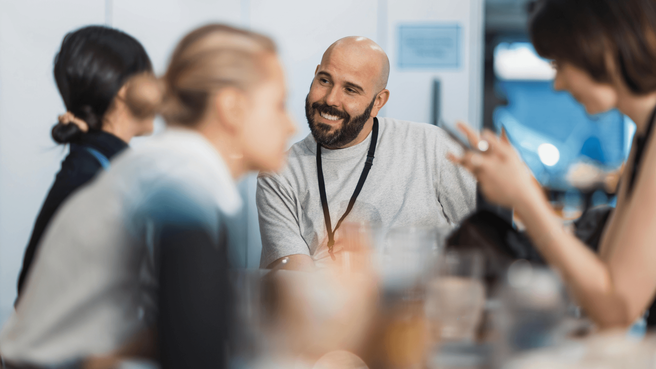 Group of people sitting and chatting, with a smiling bald man wearing a lanyard in focus, in a casual setting.