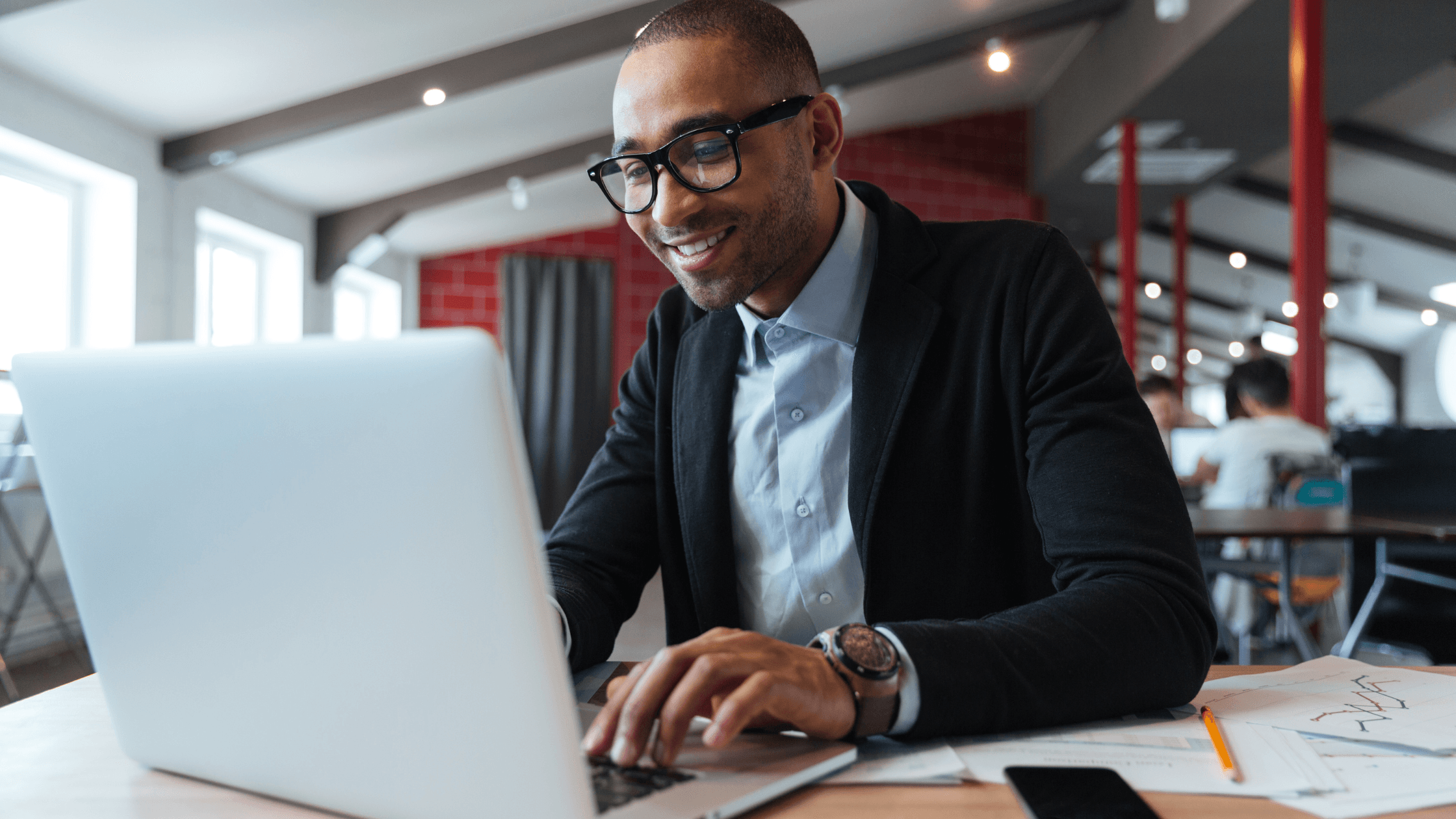 Smiling man in glasses works on a laptop in a modern office space, surrounded by charts and a smartphone.