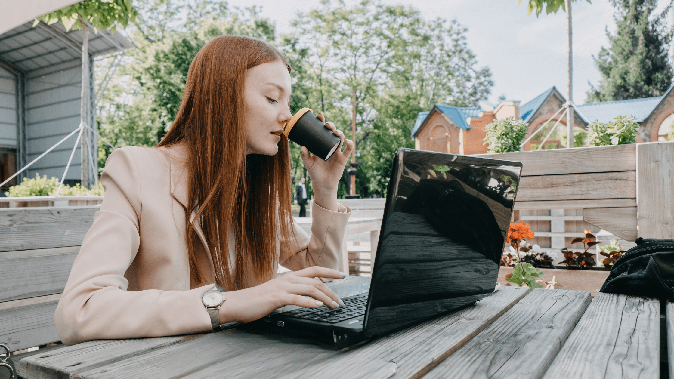 Woman with long red hair working on a laptop outdoors, sipping from a cup, seated at a wooden table surrounded by greenery and flowers.