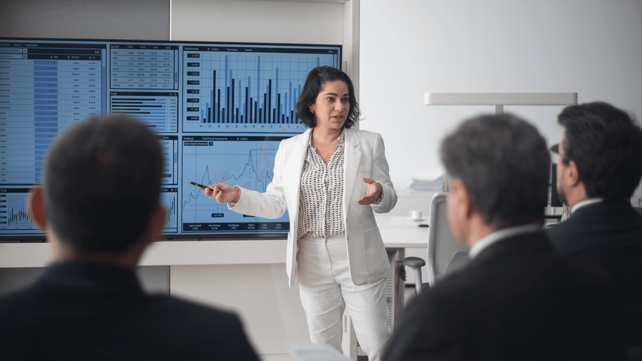 A woman in a white suit presents data on large screens to a group of seated people in a modern conference room.