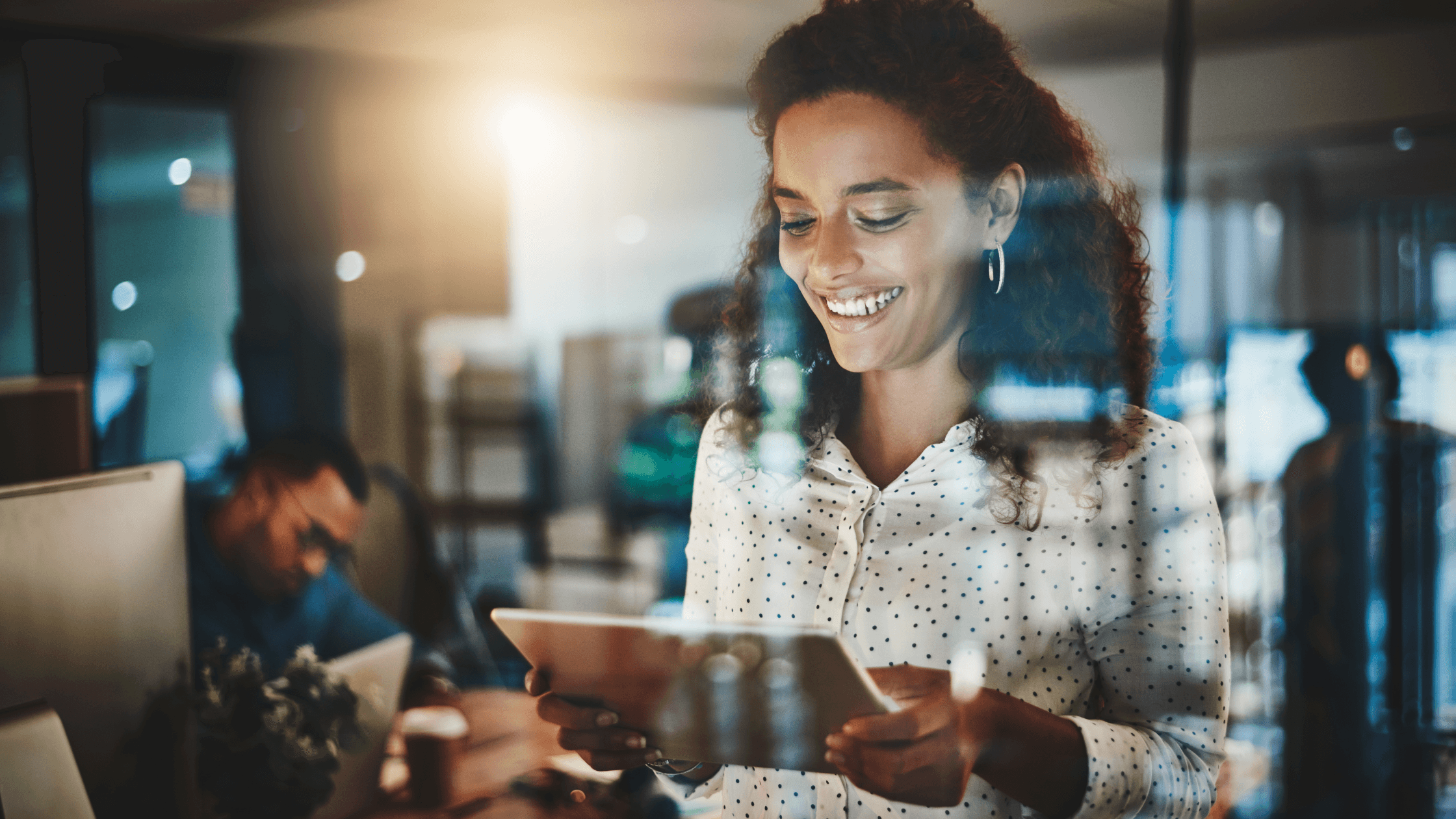 Smiling woman with curly hair using a tablet in a modern office, with another person working in the background and a warm light illuminating the scene.