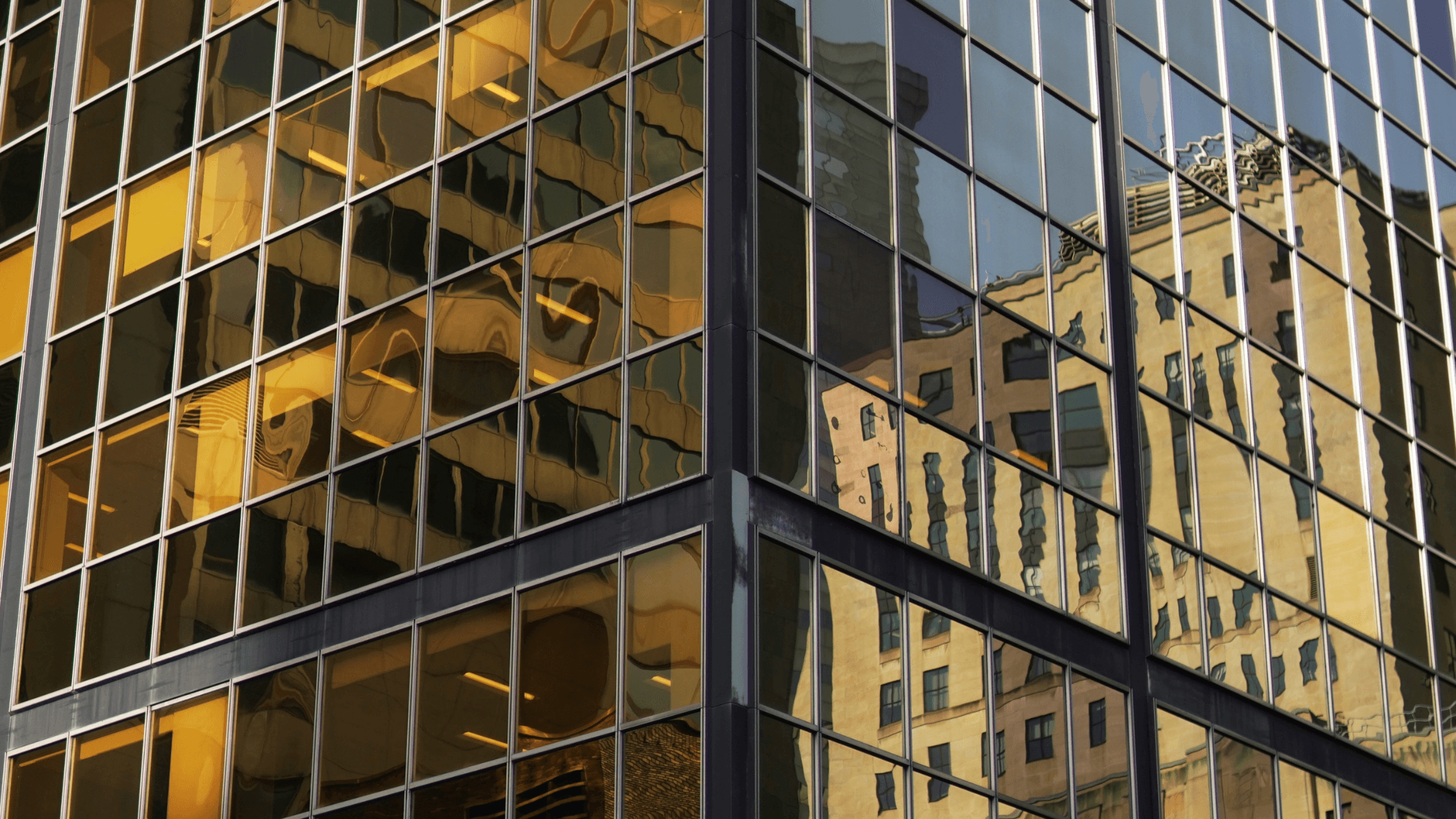Reflective glass skyscraper corner with distorted reflections of nearby buildings, creating a mosaic of urban architecture in warm tones.