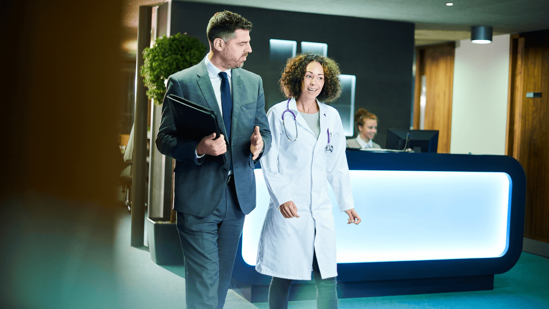 A doctor and a businessman walk through a modern office reception area, engaged in conversation, with a receptionist working in the background.