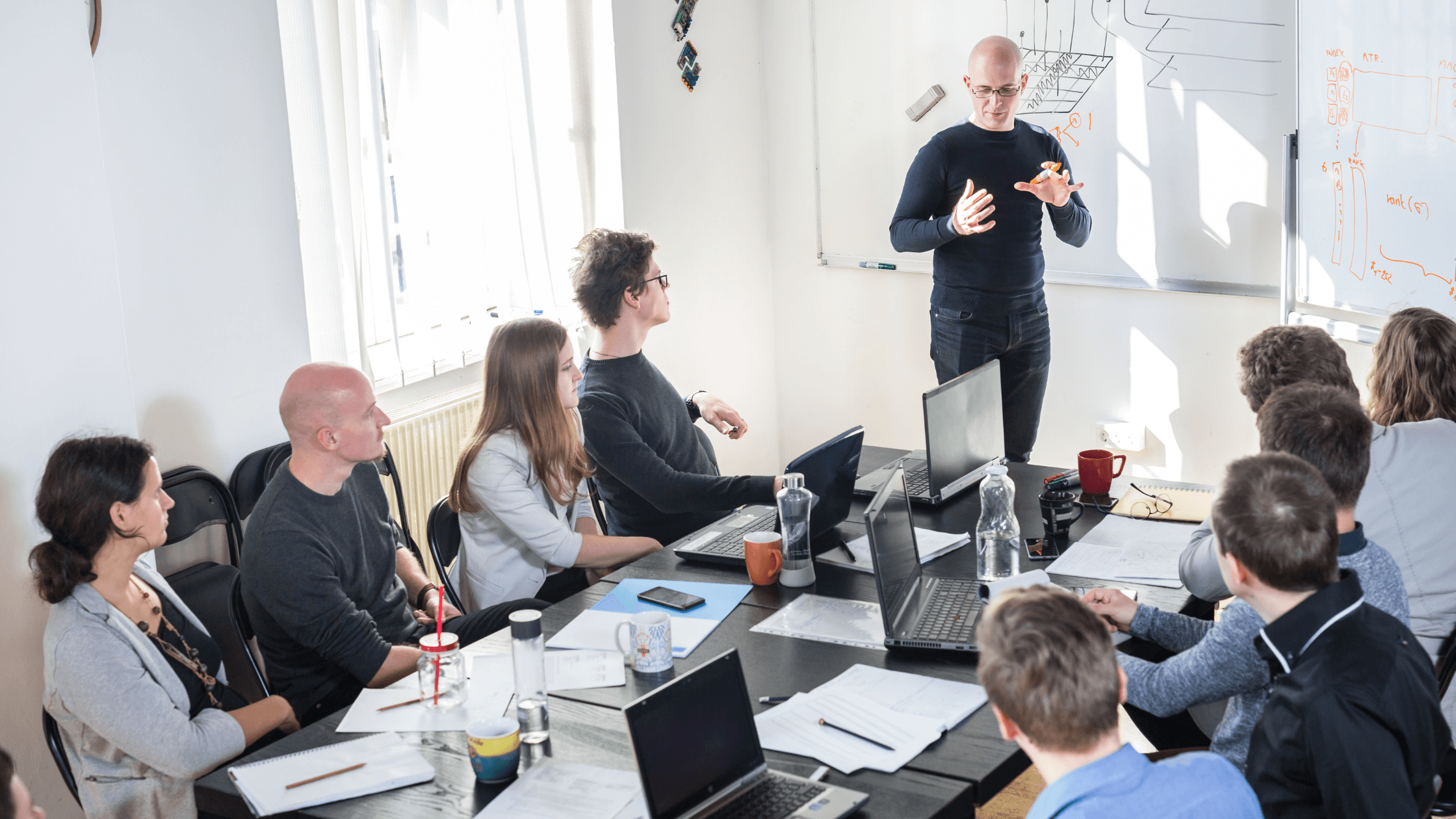 A man presents to a group of colleagues seated around a table with laptops and documents in a bright meeting room.