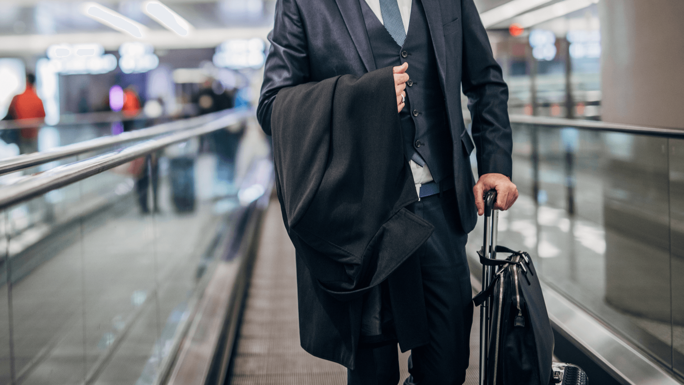Man in a suit walking with a suitcase on an airport moving walkway, holding a coat over his arm.
