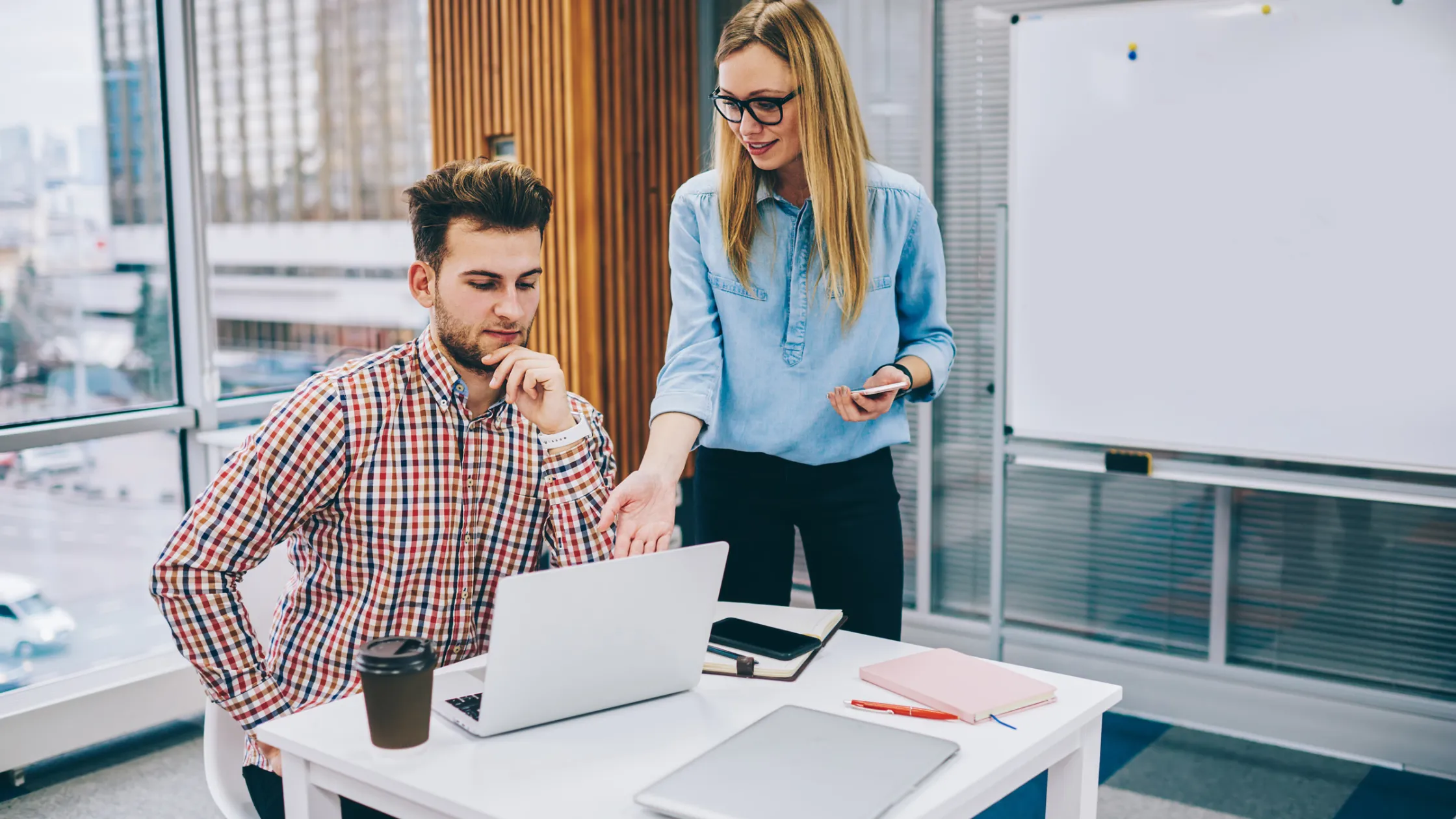 A woman in a denim shirt explains something to a man in a plaid shirt sitting at a desk with a laptop, notebooks, and a coffee cup.