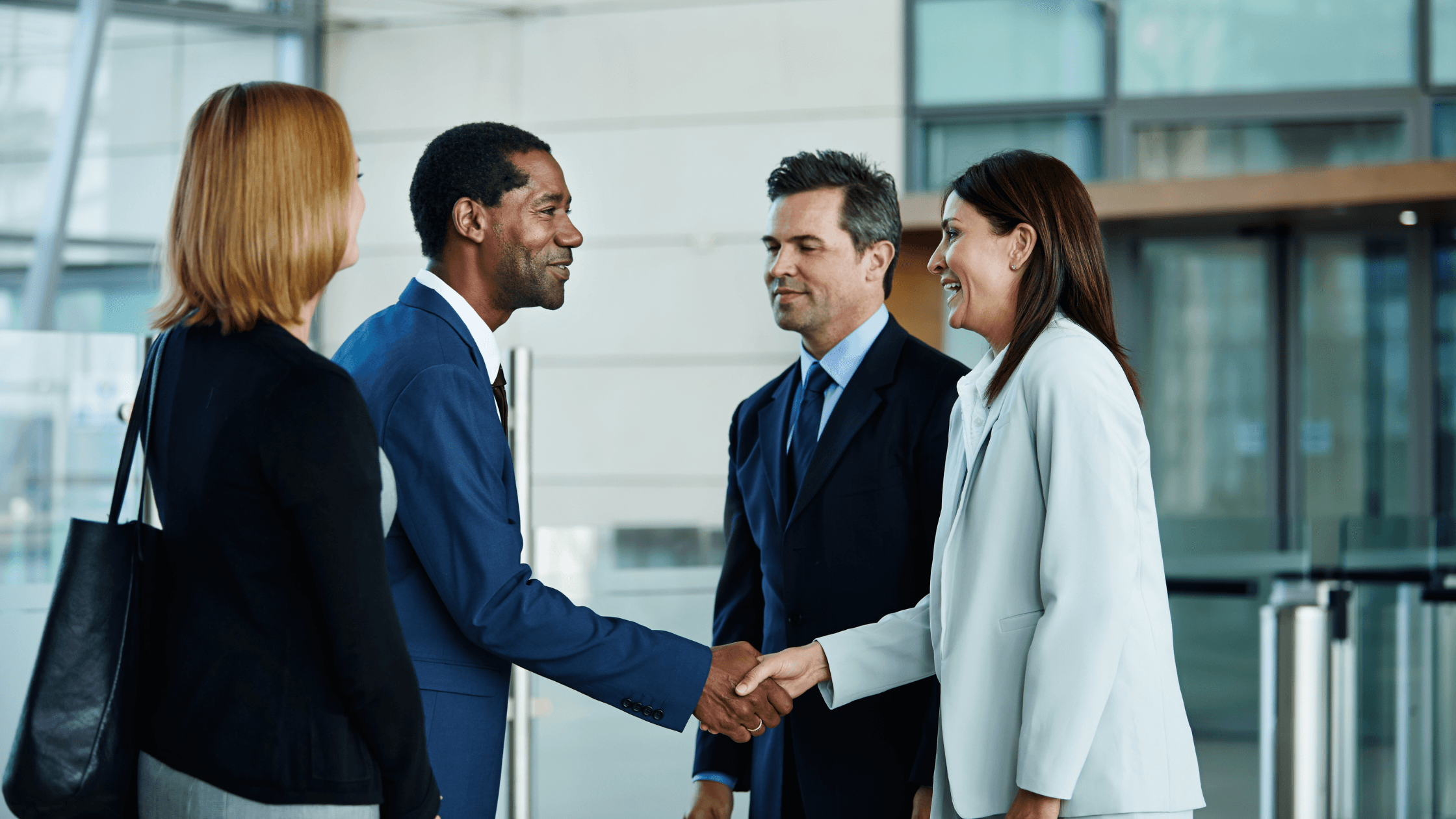 Four business professionals in formal attire are standing in a modern office lobby. Two are shaking hands, smiling, while the others look on.