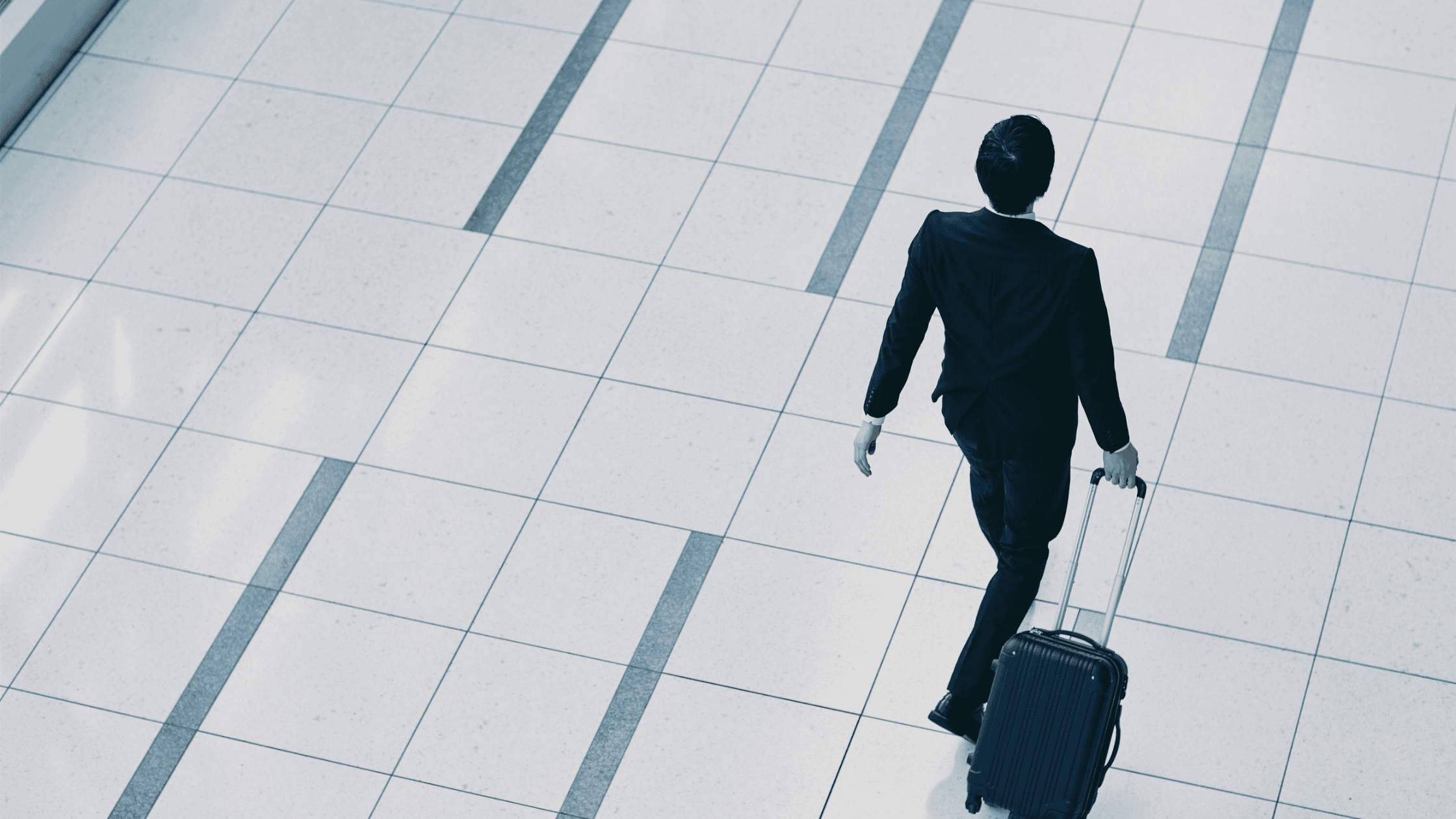Person in a suit walking on a tiled floor, pulling a suitcase. The scene is viewed from above, showing a sense of motion and travel.