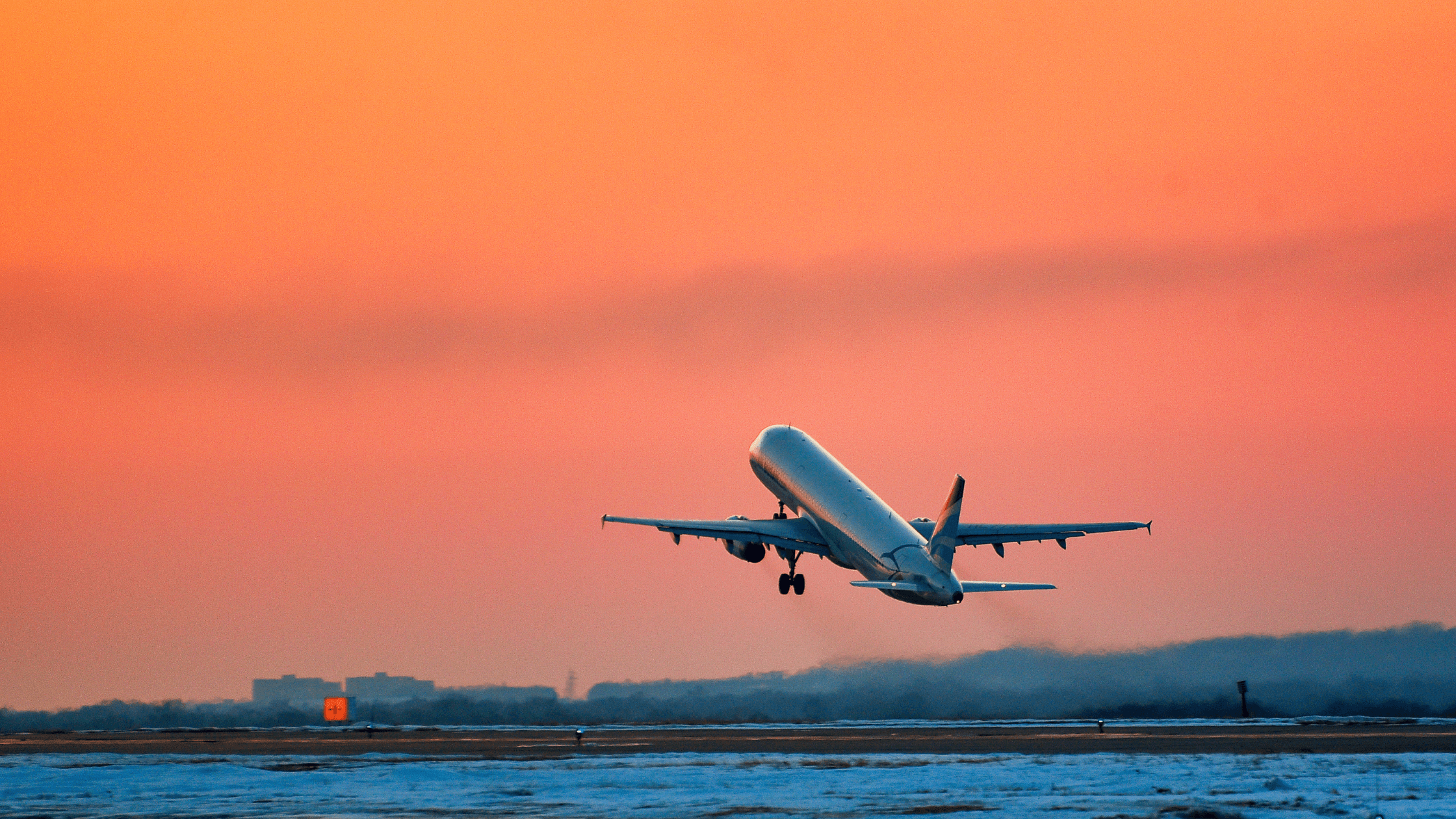 Airplane taking off against a vibrant orange sunset sky, flying over a snowy runway.