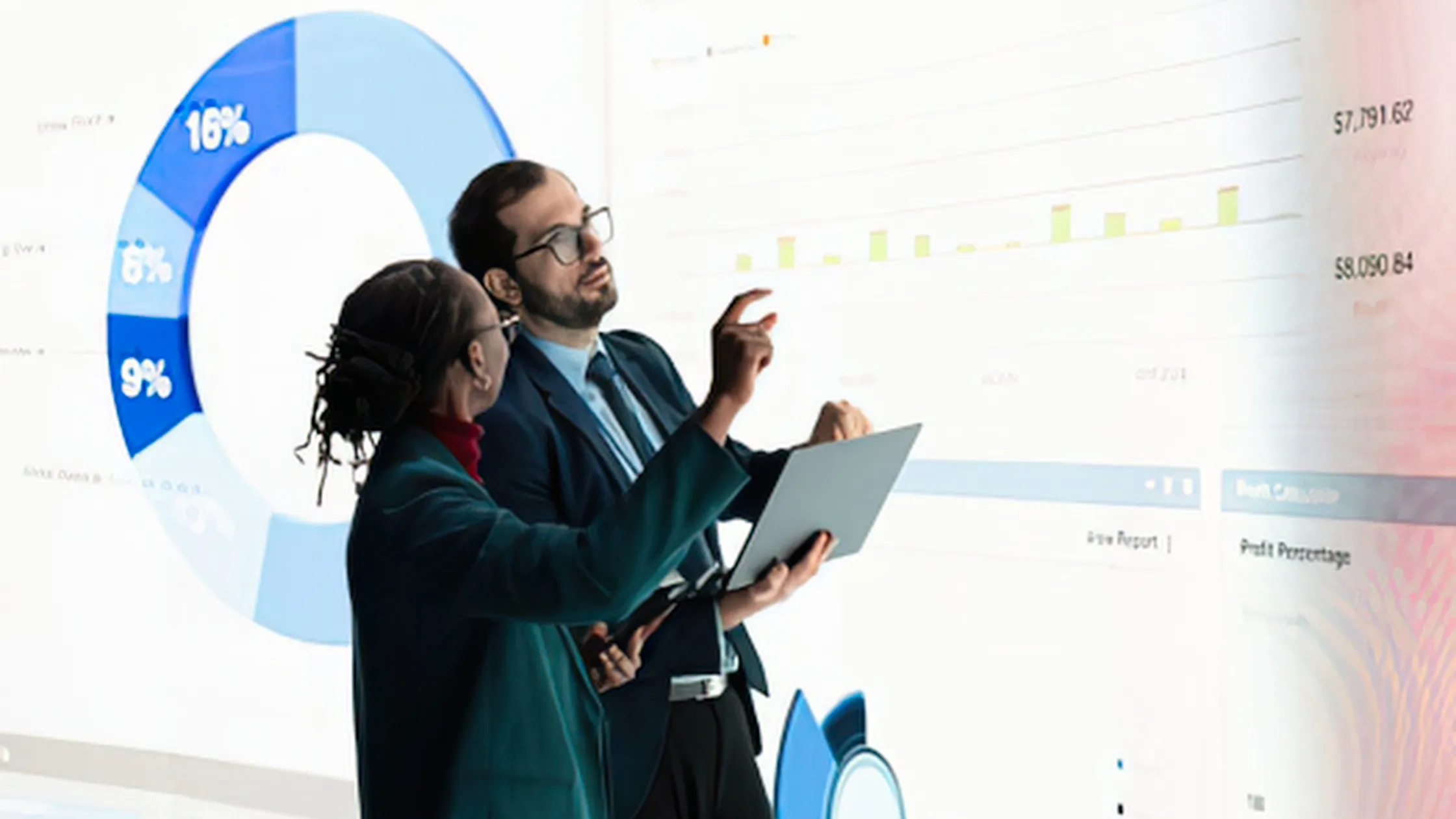 Two professionals discuss data charts on a large screen; one points at graphs while holding a tablet, both in business attire.