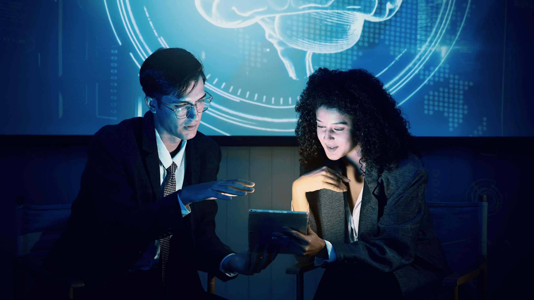 Two people in suits examine a tablet. A digital brain projection appears behind them, surrounded by data graphics, in a dimly lit setting.