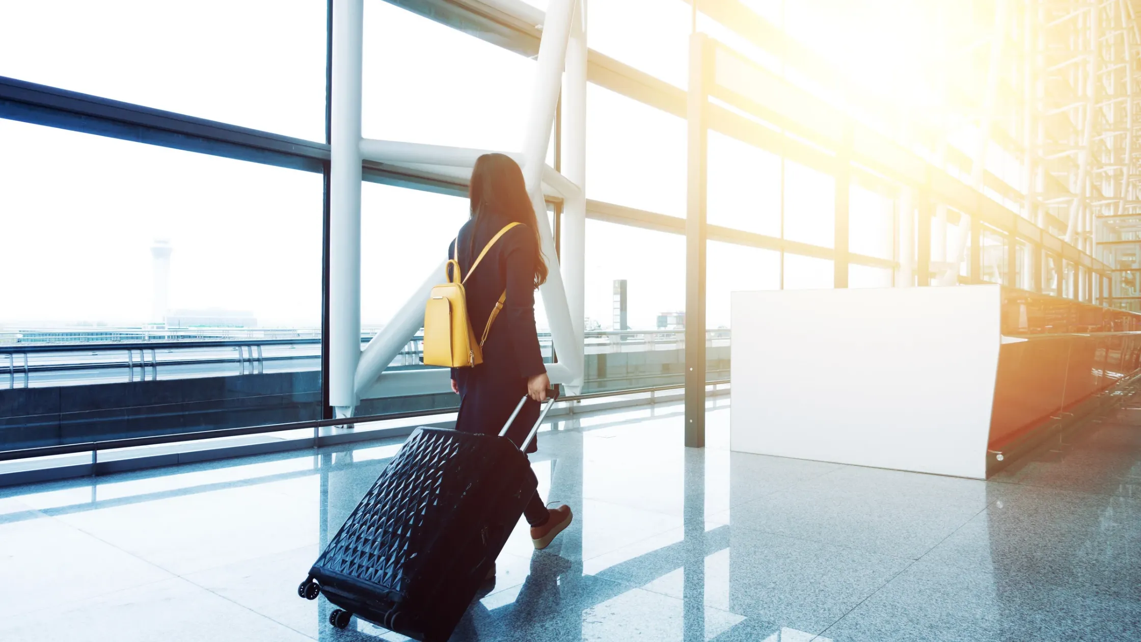 Person walking with a suitcase and yellow backpack in a sunlit airport terminal.