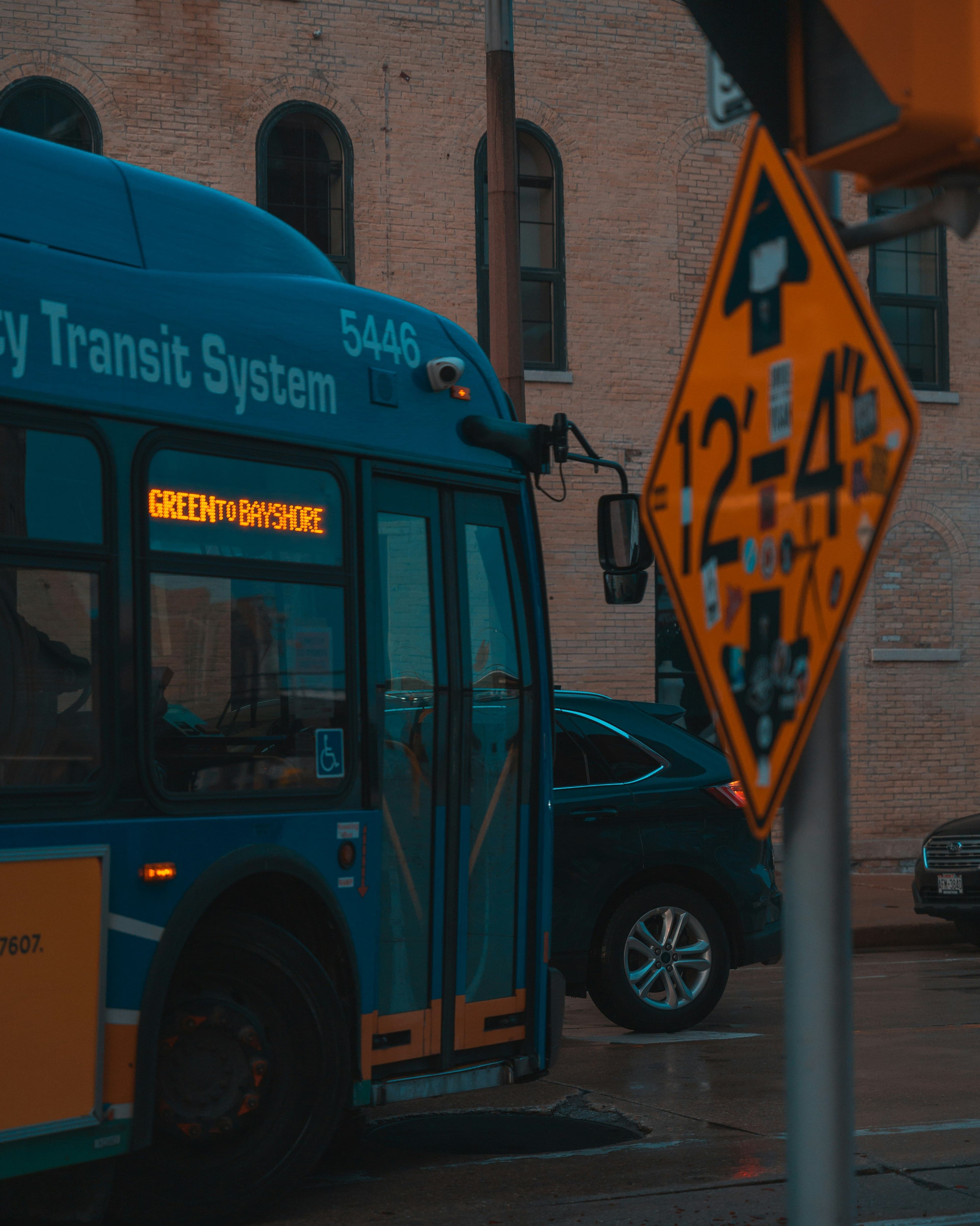 Blue city bus with "Green to Bayshore" on display, passing a yellow clearance sign and parked cars, in an urban setting with brick buildings.