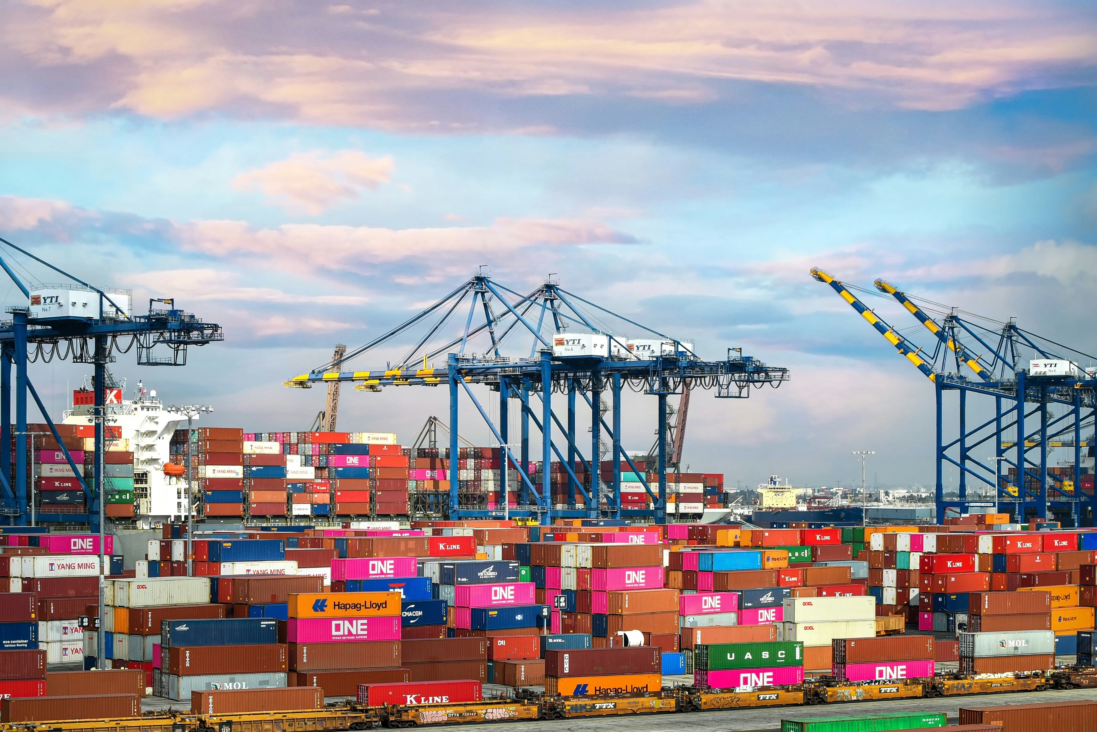 A busy shipping port with stacked colorful containers, large cranes, and a cloudy sky in the background.