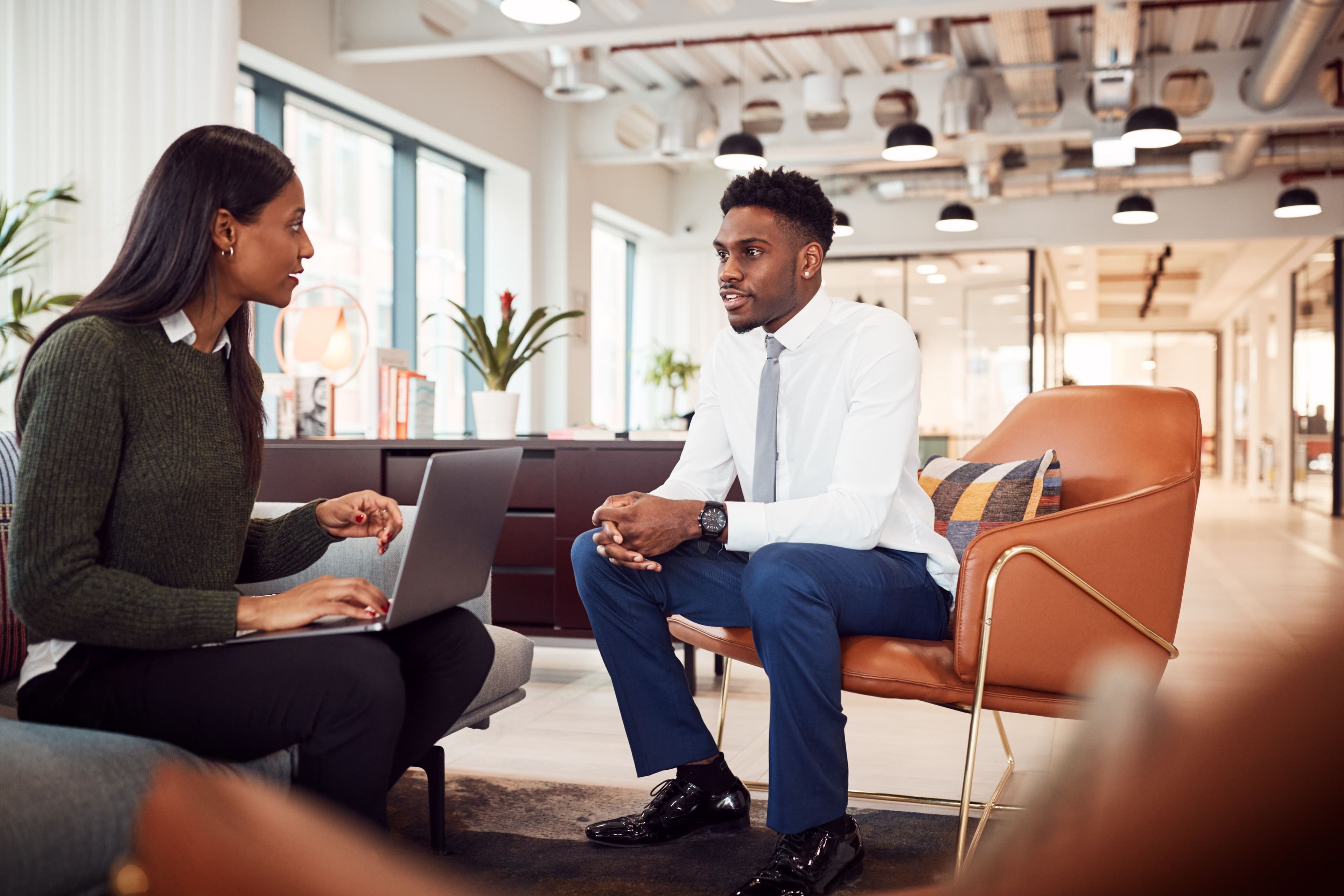 Two professionals in a modern office, one is a talent acquisition professional using a laptop while conversing with the other, who is seated and listening attentively interviewing for the job.