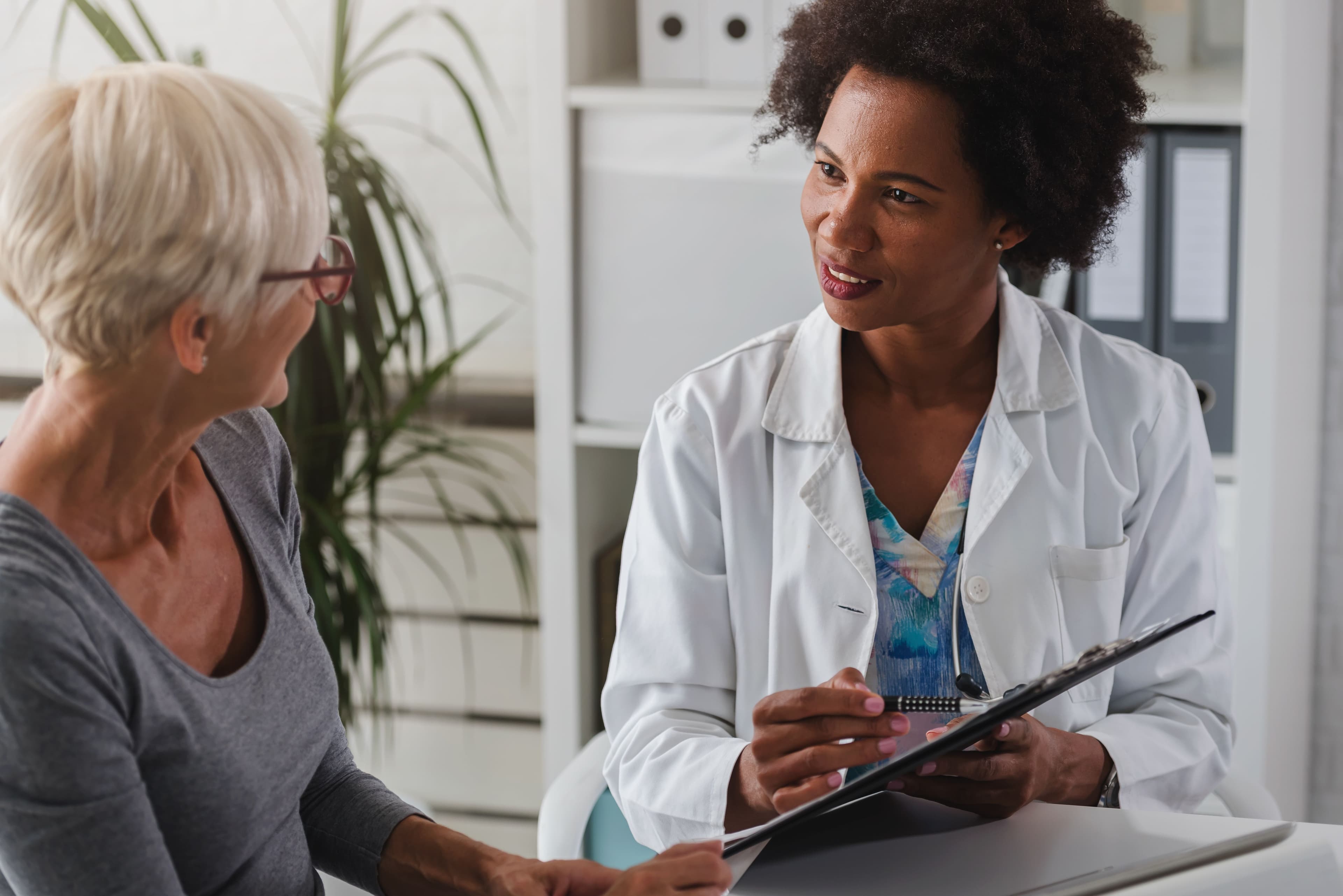 Doctor in a white coat discussing with an older woman in an office setting, holding a clipboard.