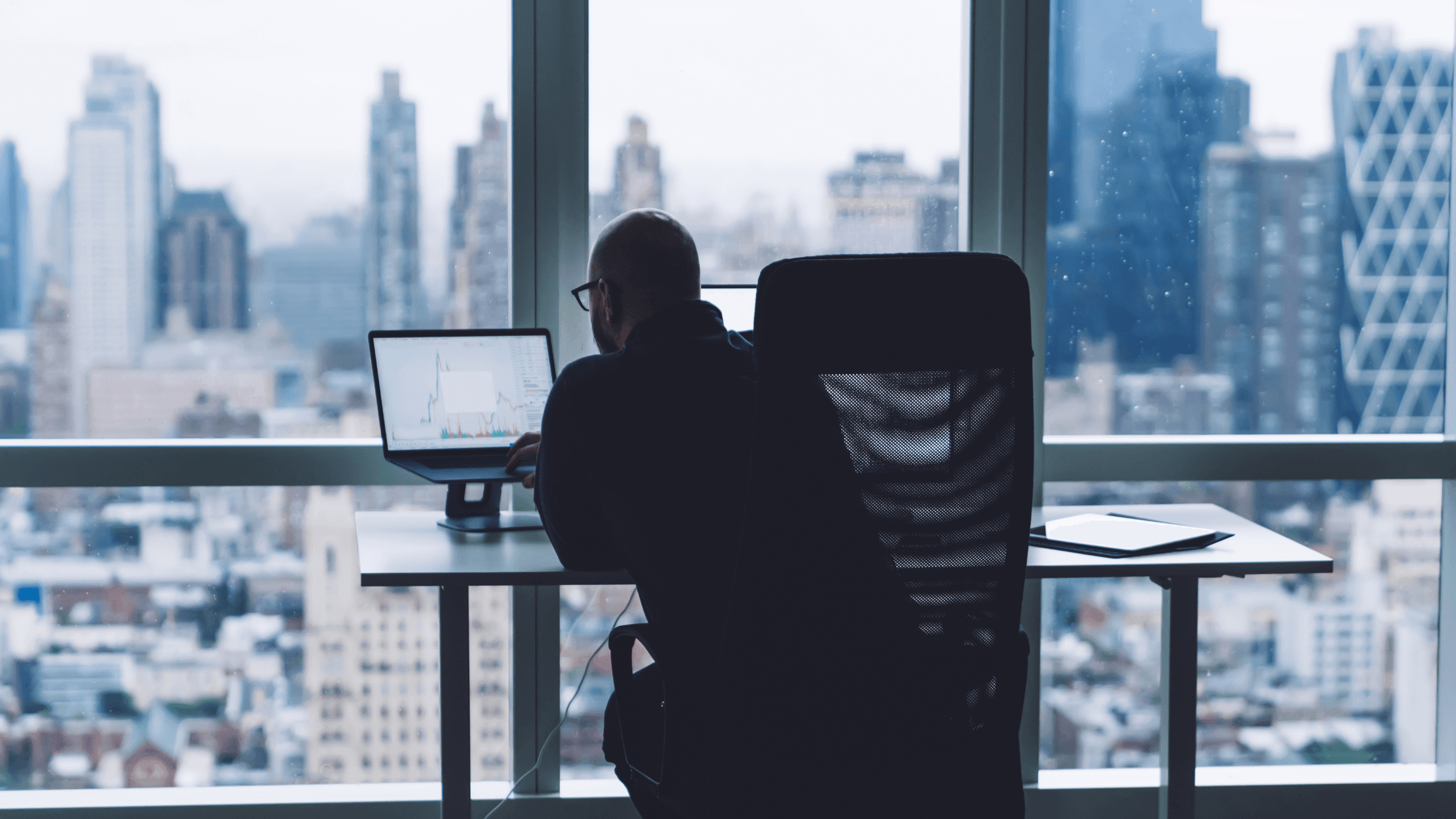Person working at a desk with a computer, overlooking a city skyline through large windows.