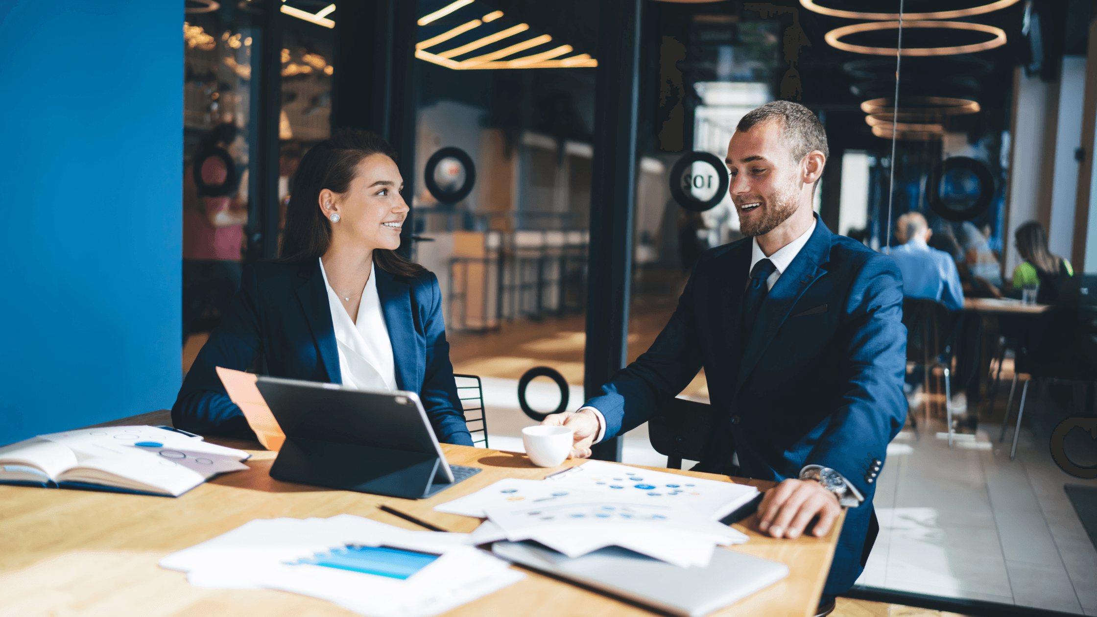 Two business professionals in suits having a meeting at a table with documents, a tablet, and a cup of coffee in a modern office setting.