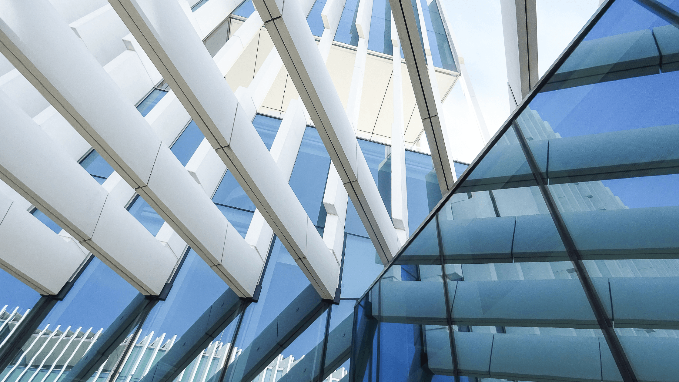 Modern building exterior featuring angled glass panels and vertical white beams with a clear blue sky in the background.