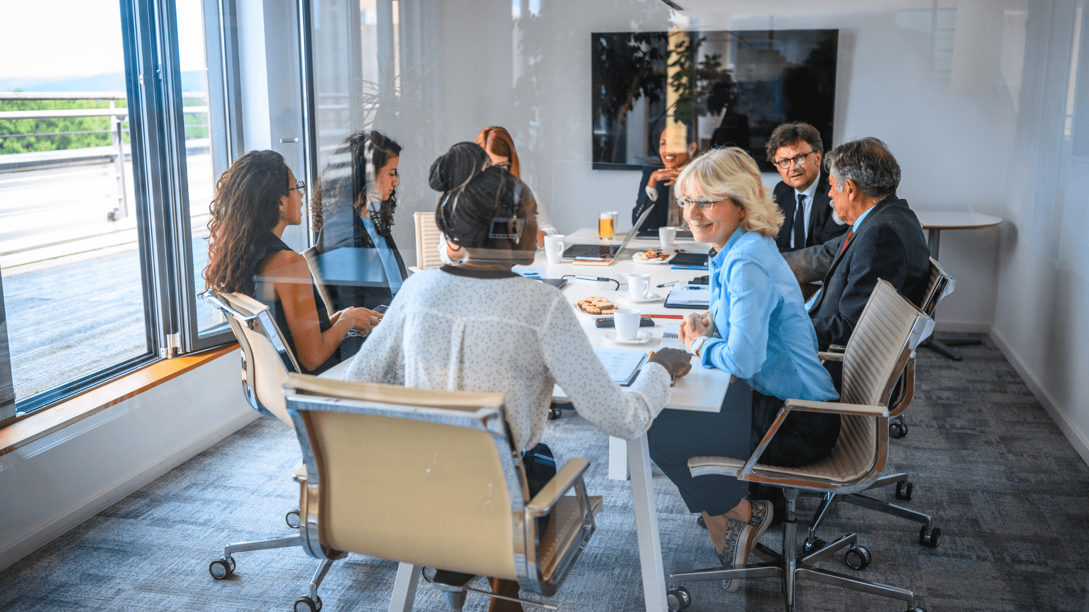 A diverse group of professionals engaged in a meeting around a conference table in a modern office with large windows.