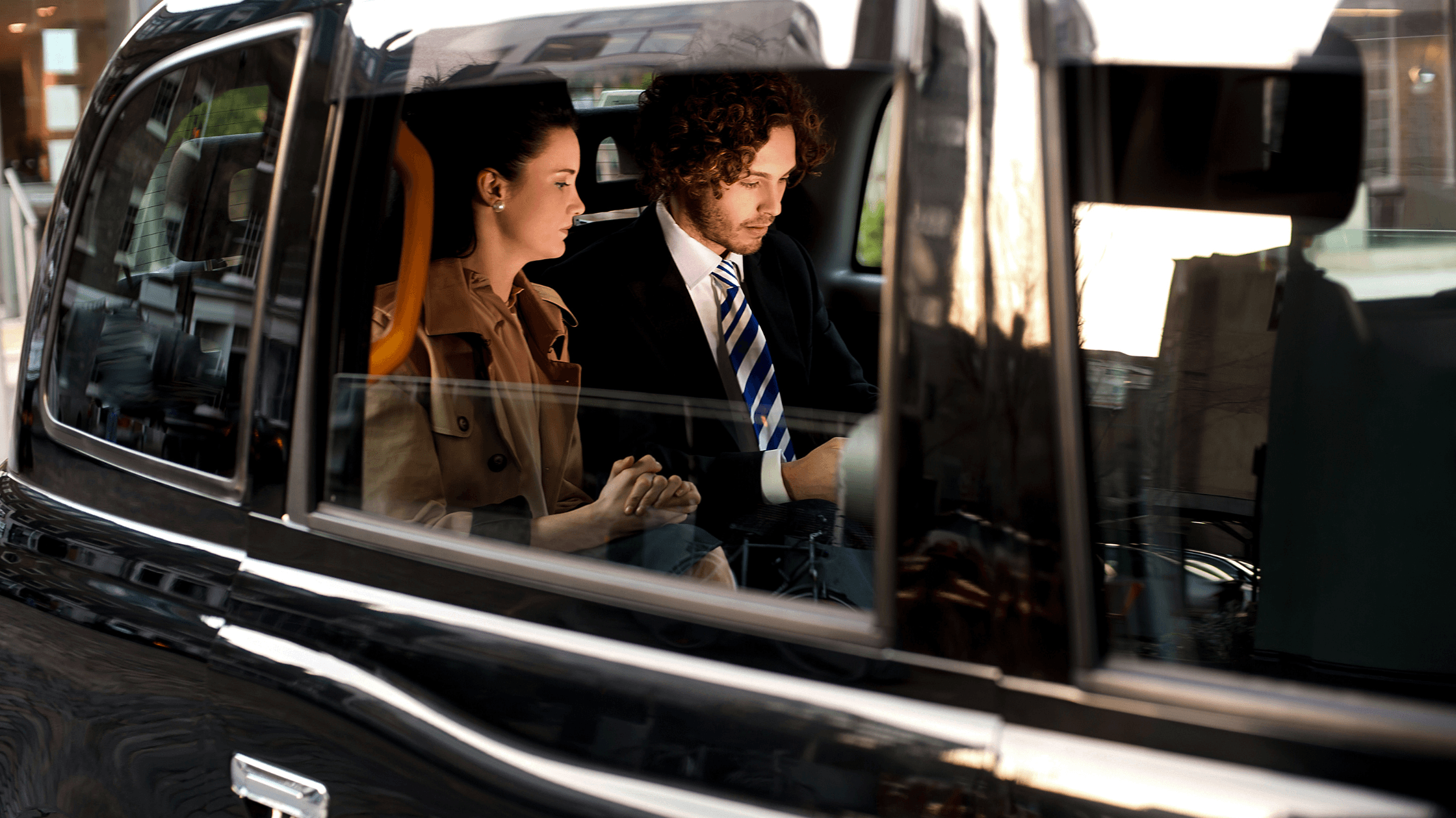 Two people sit inside a black car, one wearing a trench coat and the other a suit with a striped tie, both looking focused.