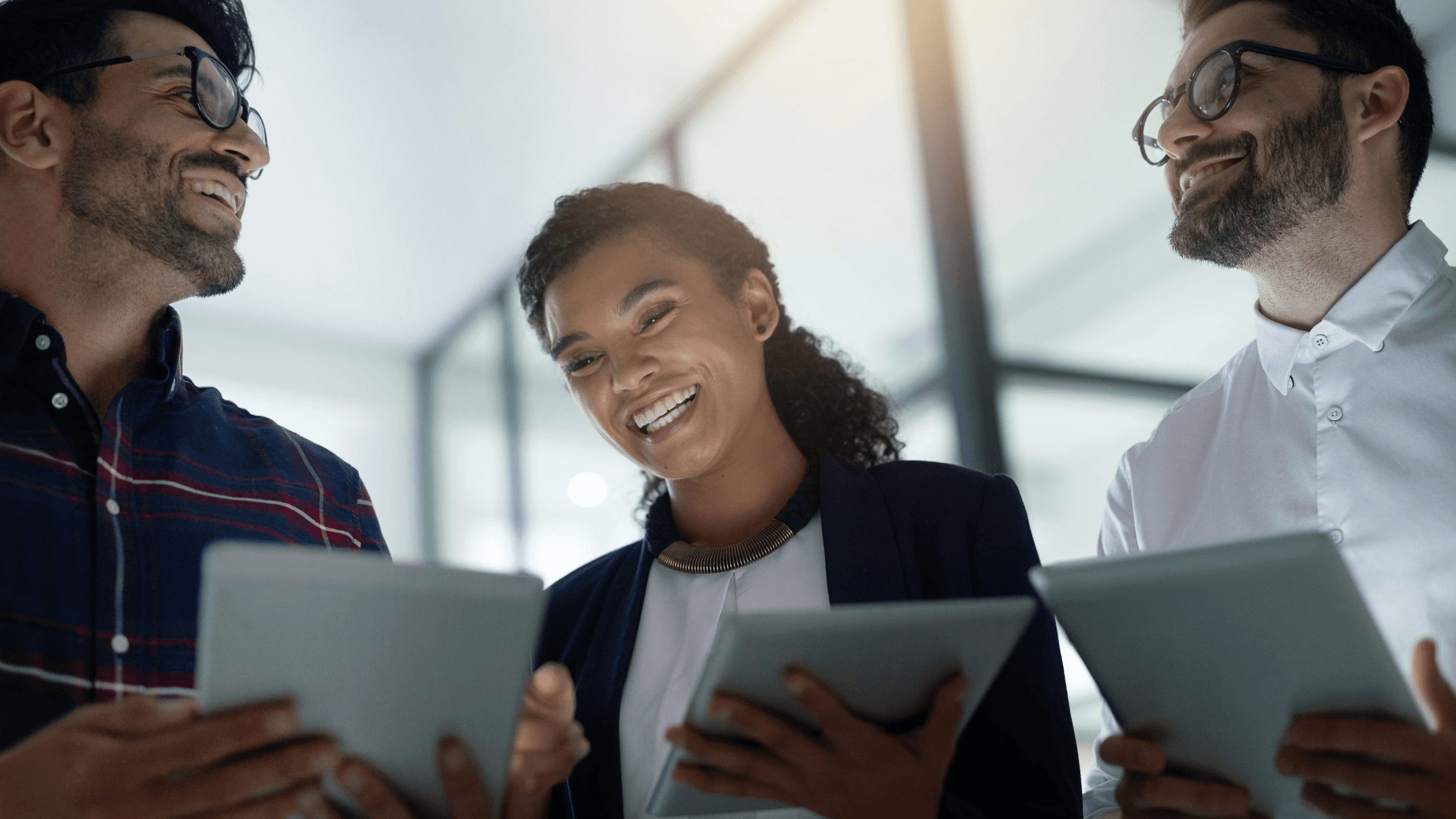 Three people smiling, standing together indoors, holding tablets. The setting appears to be a modern office with glass partitions.