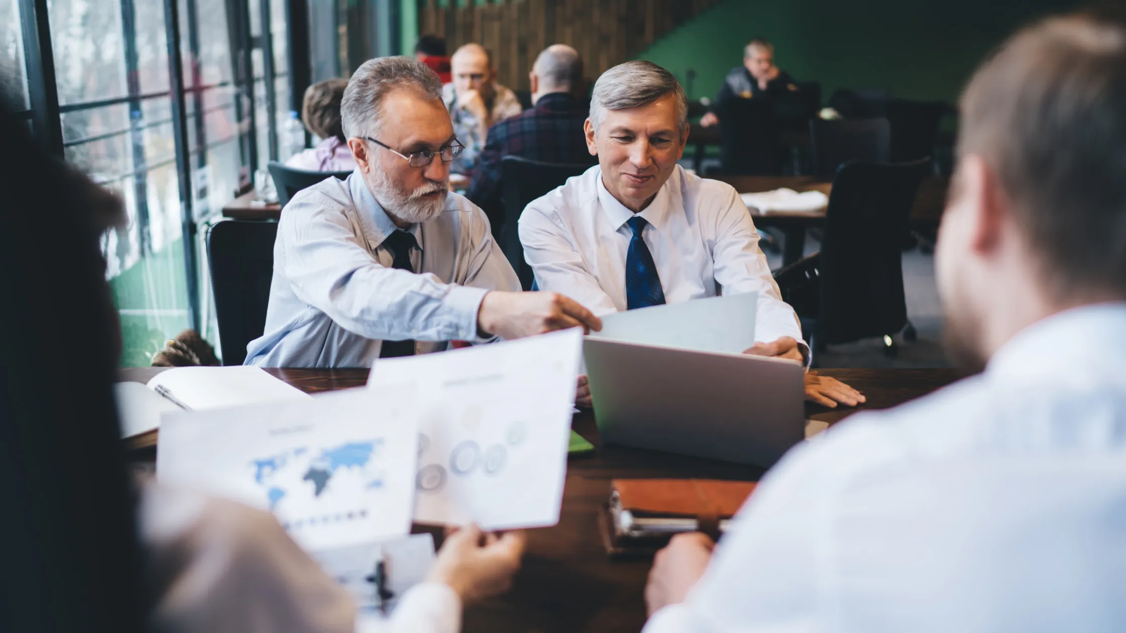 Two men in formal attire discuss documents and charts at a table in an office setting with other people in the background.