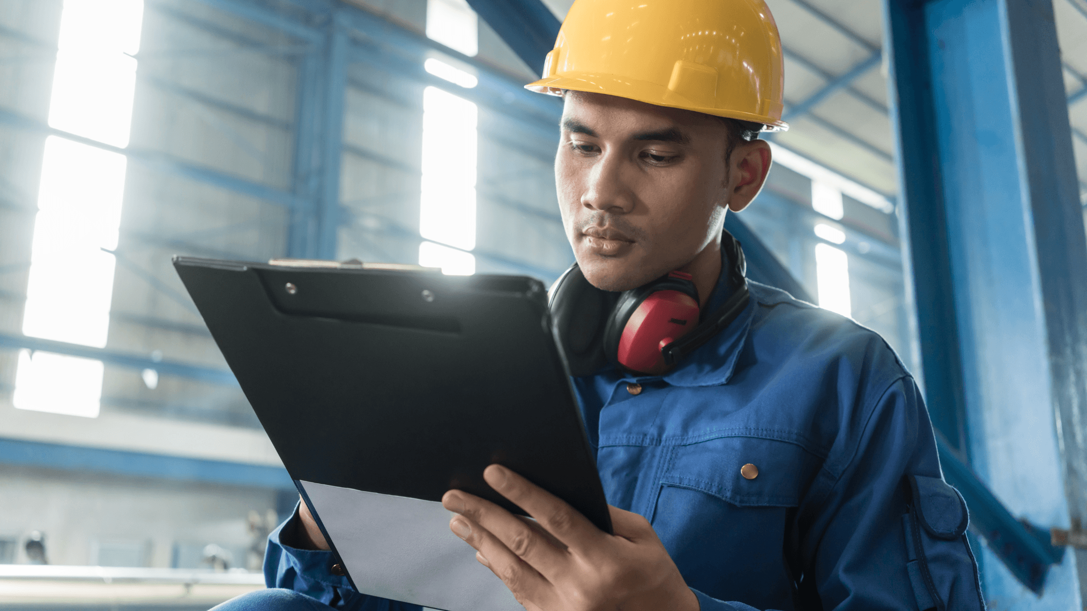 Worker in a blue uniform and yellow hard hat holding a clipboard, focused, in an industrial setting with ear protection around his neck.