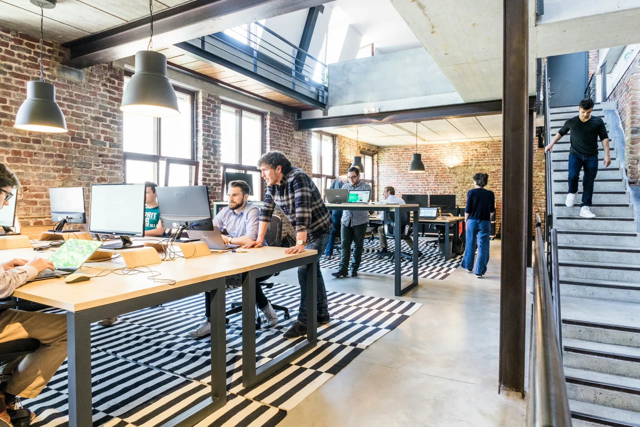 Open-plan office with people working at desks and computers. Exposed brick walls, striped rugs, and a person descending stairs on the right.
