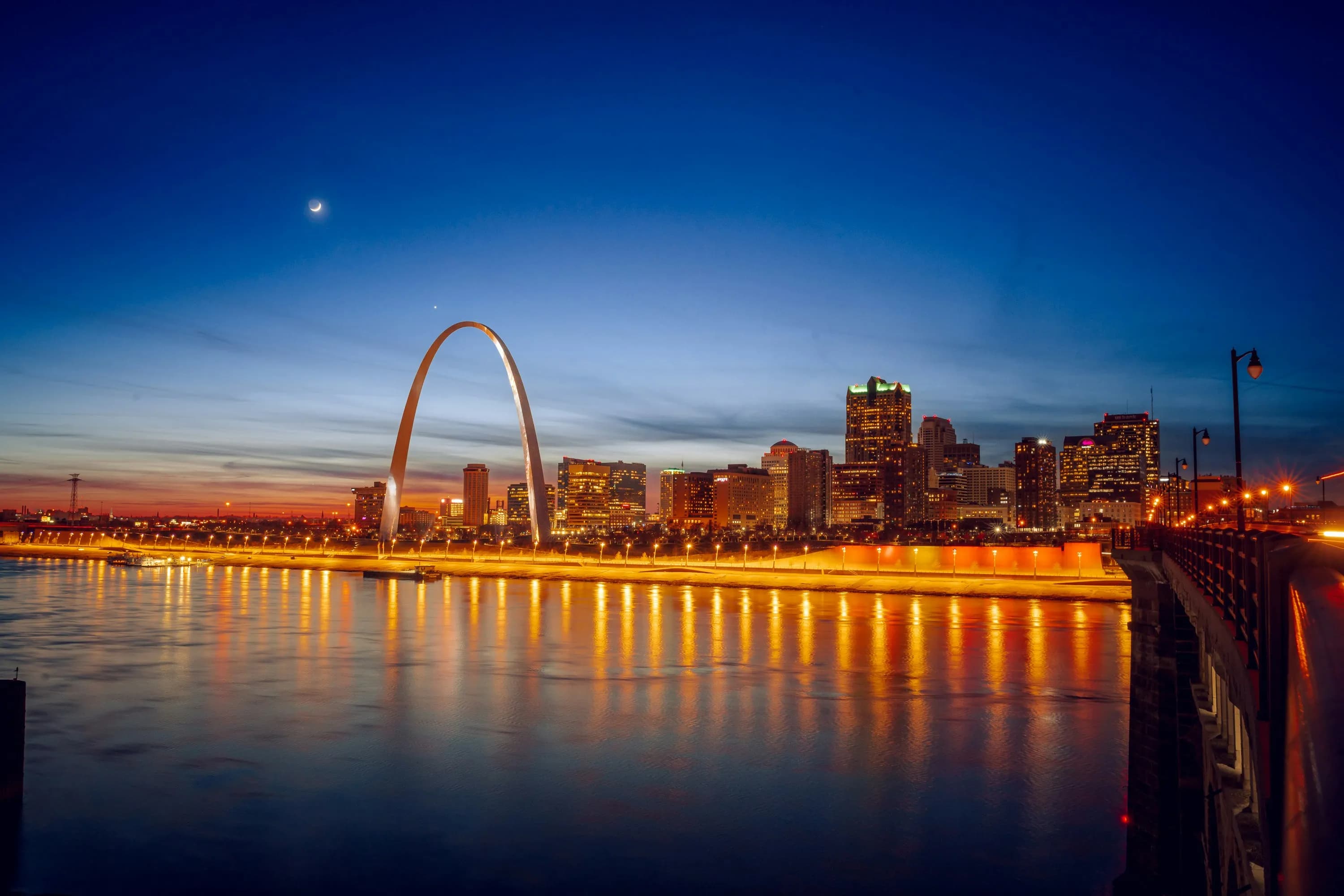 City skyline at dusk with a crescent moon, featuring an illuminated arch and reflection on the river.