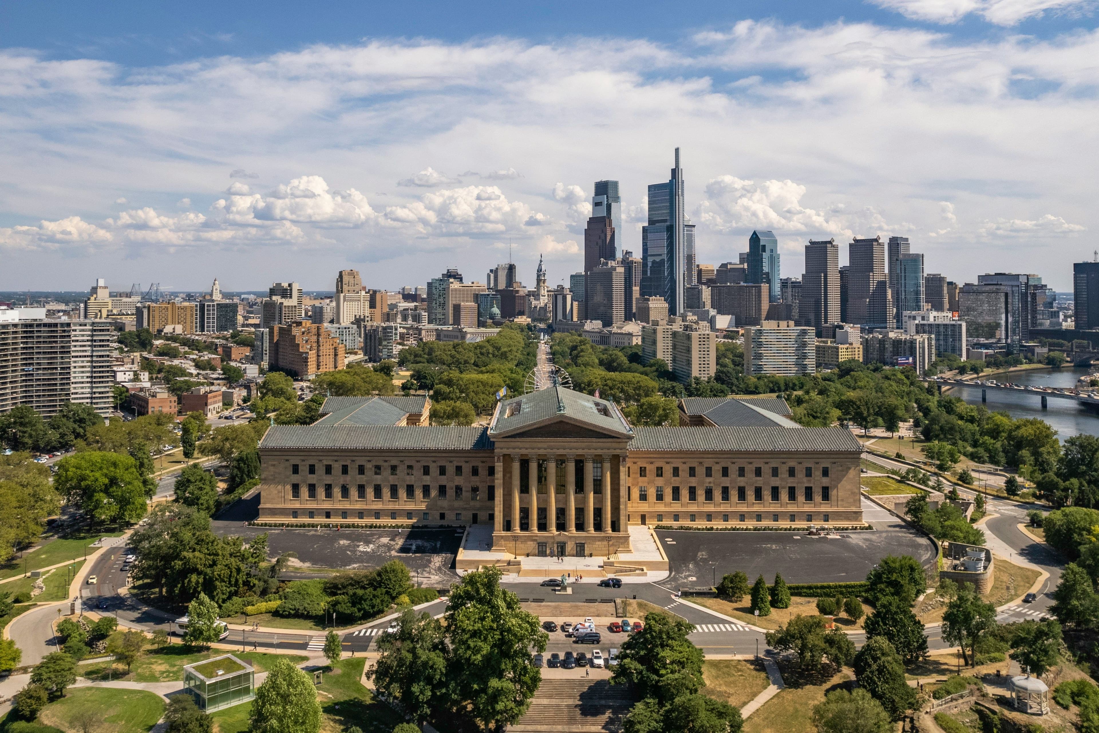 Aerial view of a grand neoclassical building with columns, set against a bustling city skyline under a partly cloudy blue sky.
