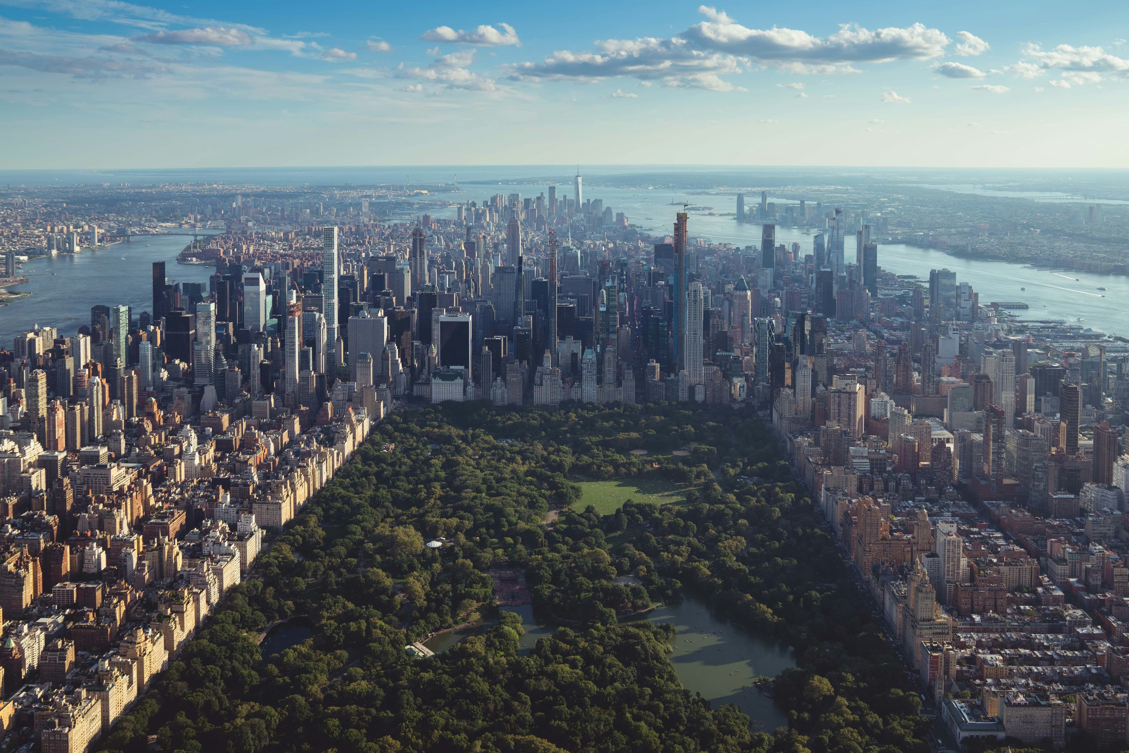 Aerial view of a large cityscape with a prominent park in the center, surrounded by tall skyscrapers and bordered by two rivers under a blue sky.
