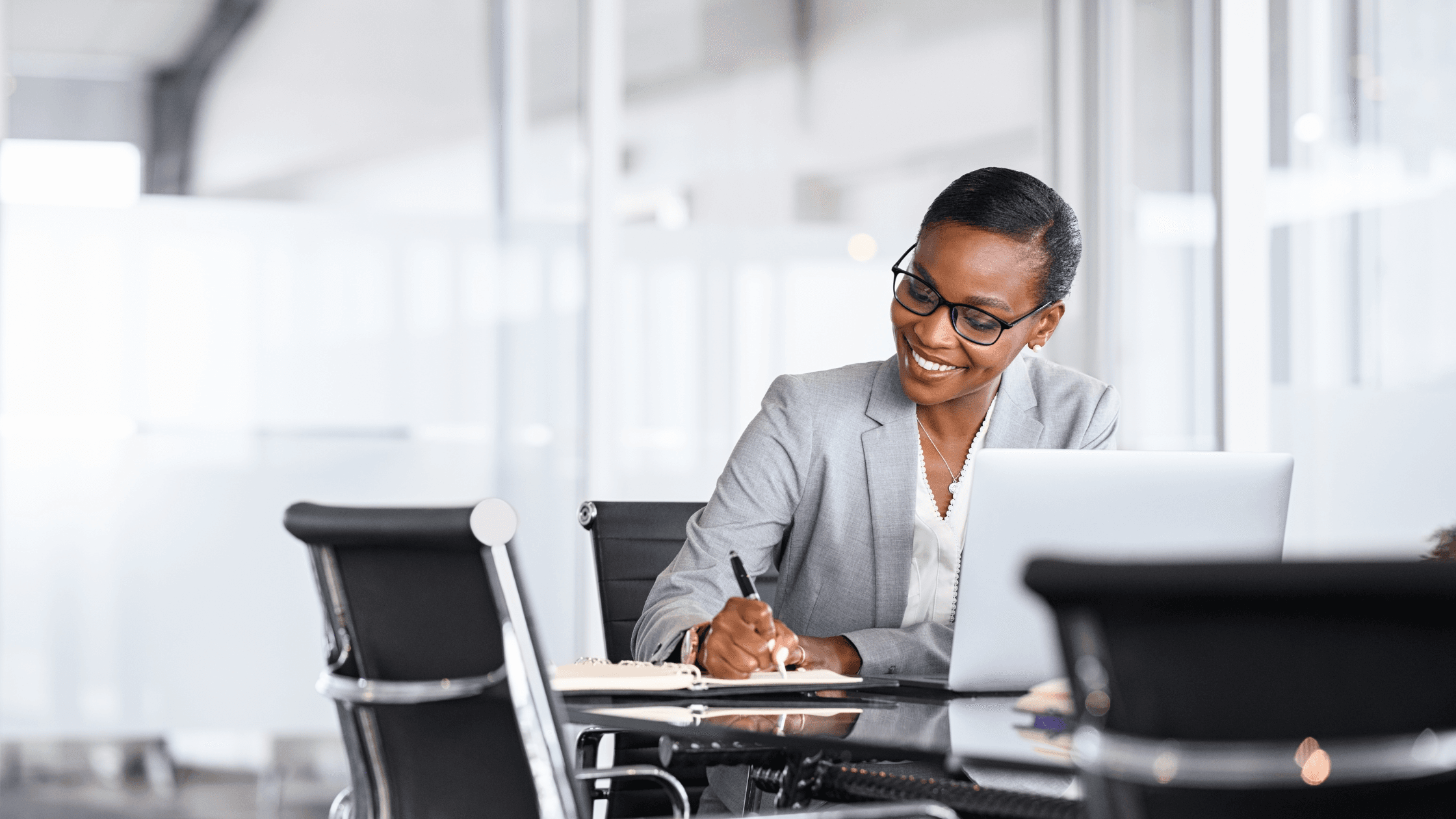 A businesswoman in a gray suit sits at a desk, smiling while writing in a notebook, with a laptop open in front of her in a modern office setting.