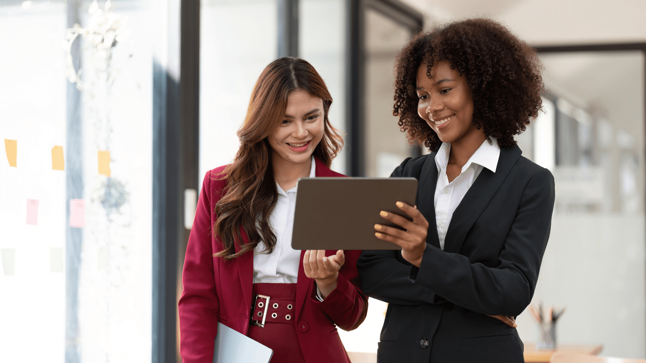 Two women in business attire smiling while looking at a tablet in a bright office setting.